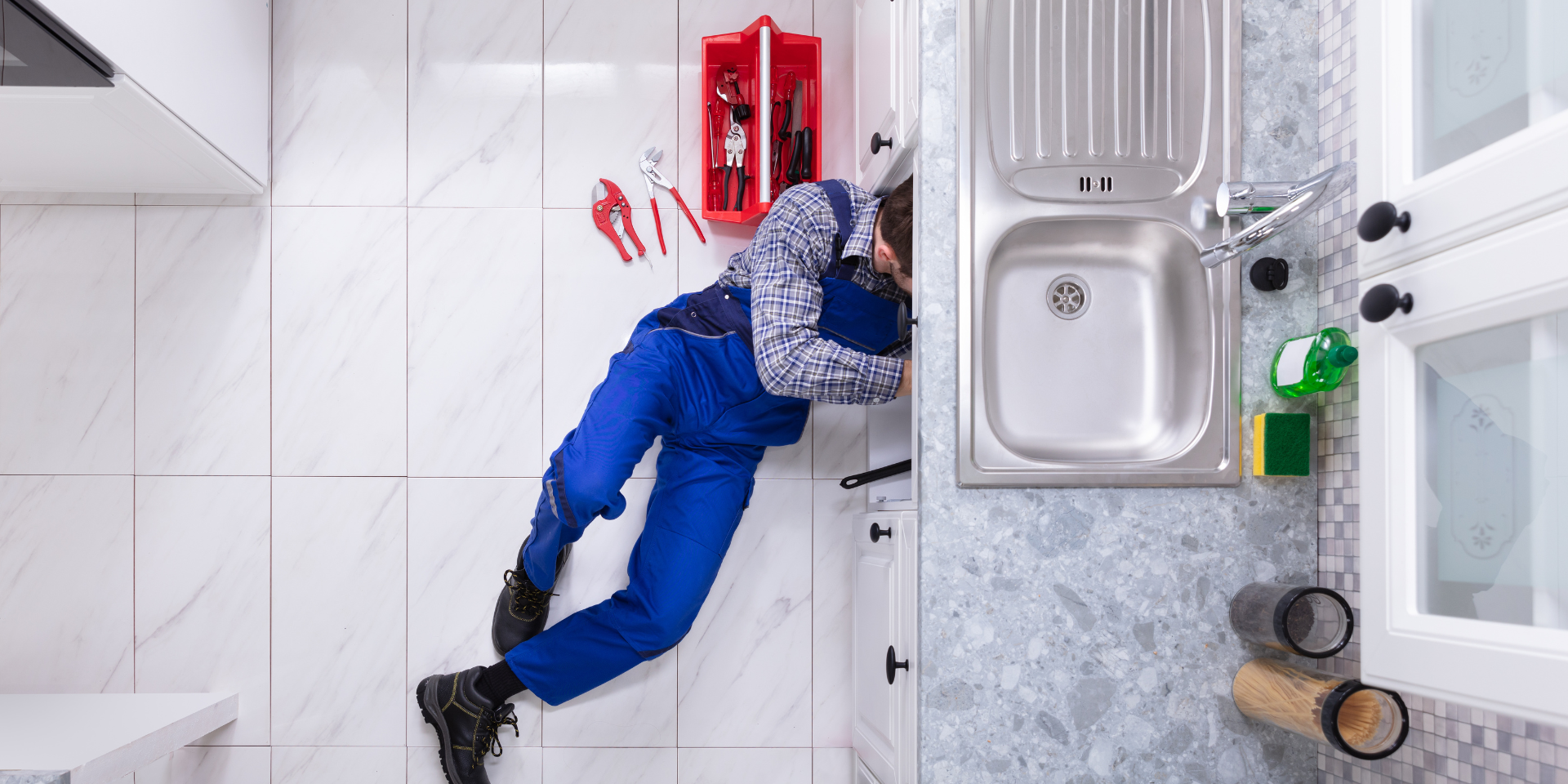 Un plombier en salopette bleue répare un tuyau sous un évier de cuisine, allongé sur le sol en carrelage blanc. Une boîte à outils rouge et plusieurs pinces sont disposées à côté de lui.