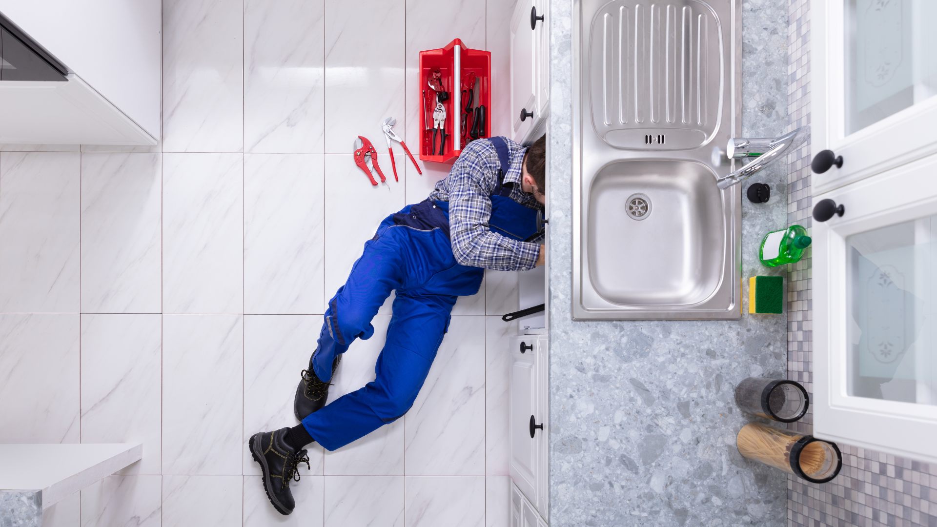 Un plombier en salopette bleue répare un tuyau sous un évier de cuisine, allongé sur le sol en carrelage blanc. Une boîte à outils rouge et plusieurs pinces sont disposées à côté de lui.